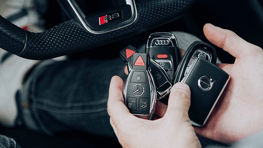 Zwei Hände halten verschiedene Autoschlüssel (Mercedes, Nissan, Audi, zwei nicht erkennbar). Im Hintergrund ist ein Lenkrad zu erkennen